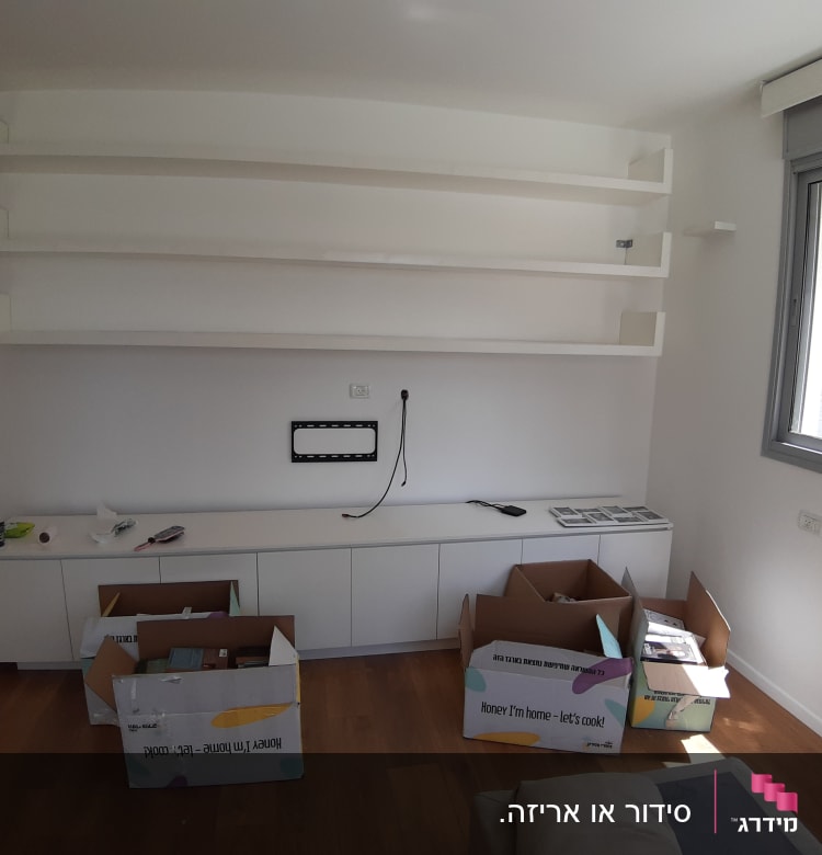 קופסאות קרטון פתוחות על רצפת עץ ליד מדפים לבנים